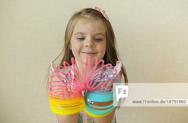 Cute adorable kleines Mädchen spielt mit Kunststoff Regenbogen magische Feder  hält bouncy dehnbar Spielzeug und lächelt in die Kamera. Glückliches Kind vorbeige Hintergrund genießen Freizeit zu Hause