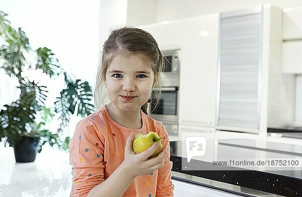 Kleines kaukasisches Mädchen ißt einen großen roten Apfel in der Küche. Schönes kleines Mädchen beißt und ißt einen Apfel. Schaut in die Kamera und hält Obst. Nahaufnahme Porträt