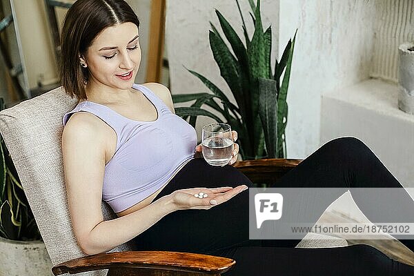 Nahrungsergänzungsmittel während der Schwangerschaft. Junge glückliche schwangere Frau  die pränatale Vitamine einnimmt  mit einem Glas Wasser und einer Pille in der Hand  während sie zu Hause im Sessel sitzt. Werdende Mutter beginnt jeden Tag mit Folsäure