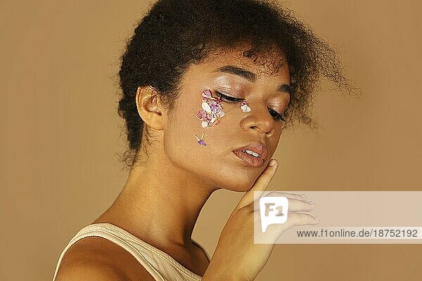 Porträt eines schönen afrikanischen Mädchens mit lockigem Haar zeigt kreatives Make up mit Blumenblättern  während die Hand sanft ihr Gesicht berührt  vorbeigem Hintergrund. Natürliche Ökokosmetik und Schönheit Konzept