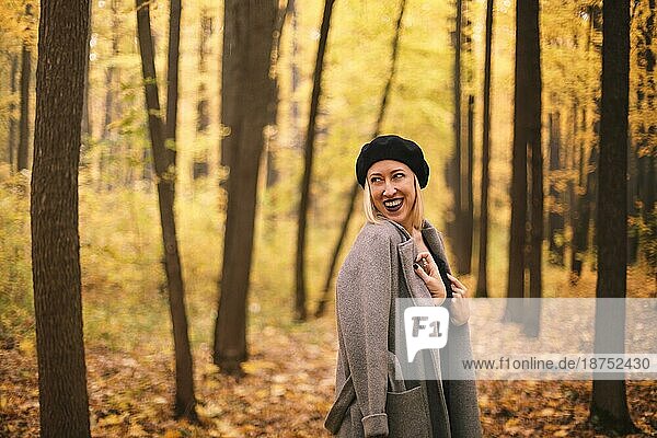 Schöne junge fröhliche europäische Frau mit Baskenmütze und Mantel spazieren im Herbst Wald  Blick weg mit glücklichen Gesichtsausdruck  glückliche Frau verbringt Zeit in der Natur auf Herbst Tag