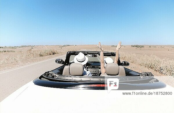 Rückenansicht eines nicht erkennbaren Paares mit Hüten  das die Freiheit genießt  während es in einem Cabrio durch eine grasbewachsene Wüstenlandschaft an einem Sommertag fährt