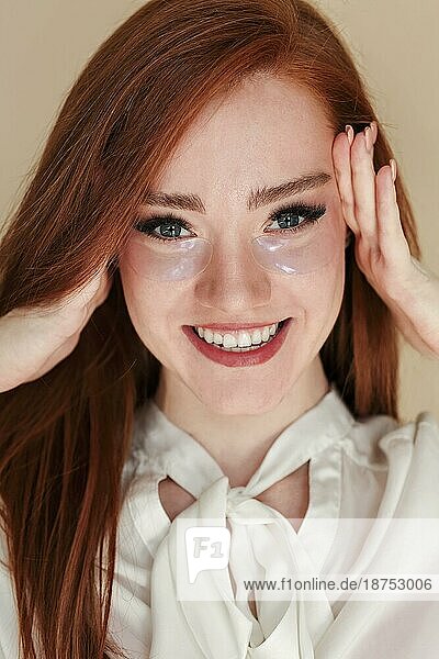 Fröhliche junge rothaarige Frau mit Hydrogel Augenklappen  die den Kopf berührt und lächelnd in die Kamera schaut  während sie ihre Haut pflegt