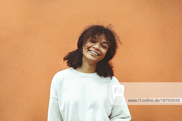 Pures Glück. Nettes glückliches afroamerikanisches Mädchen mit lockiger Frisur und weißem Pullover posiert gegen eine orangefarbene Wand. Lächelnde schwarze junge Frau  die mit ihrem Leben zufrieden ist