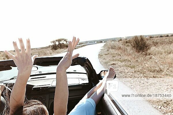 Rückenansicht einer fröhlichen jungen Frau in Sommerkleidung auf dem Beifahrersitz eines Cabriolets während einer Autofahrt