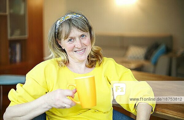 Positive Frau mittleren Alters in lässigem gelben Hemd und Jeans sitzt am Tisch mit einer Tasse Tee in der Hand und schaut in die Kamera  während sie sich zu Hause ausruht