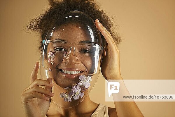 Fröhliche afroamerikanische junge Frau  die einen transparenten PVC Schutzschild trägt  der mit Blumen verziert ist und mit einem strahlenden Lächeln in die Kamera blickt  vor einer beigen Wand