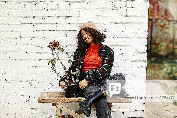 Junge  glückliche  positive  gemischtrassige Frau mit Blumentopf sitzt auf einer Holzbank in der Nähe des Hauses und lächelt  pflanzt Blumen im Garten während der Herbstzeit
