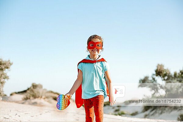 Glückliches Superheldenkind  das am Strand läuft und ein Pop It Spielzeug in der Hand hält