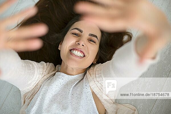 Porträt von oben einer glücklichen jungen positiven Frau  die auf einem rustikalen Holzboden liegt und beide Hände nach oben streckt und mit einem breiten  strahlenden Lächeln in die Kamera blickt  das ihre weißen  geraden  gesunden Zähne zeigt