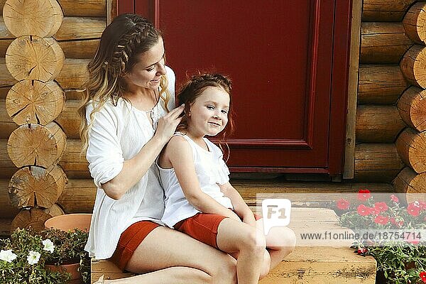 Nettes kleines rothaariges Mädchen und glückliche junge Mutter mit ähnlicher Flechtfrisur umarmen sich  während sie vor einer roten Tür stehen