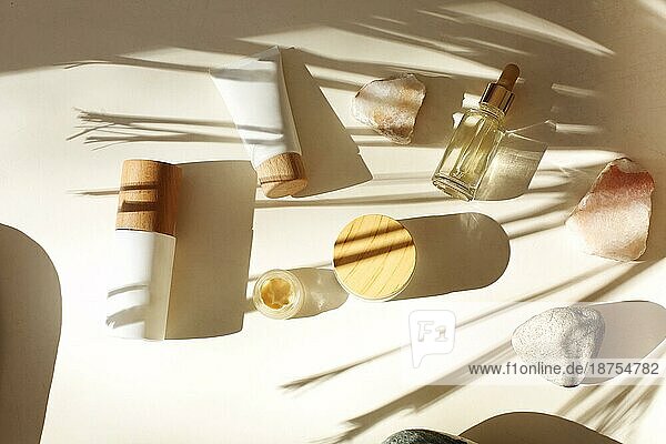 Draufsicht auf Flaschen verschiedener Kosmetikprodukte inmitten von Steinen mit Schatten einer Palme auf weißem Hintergrund