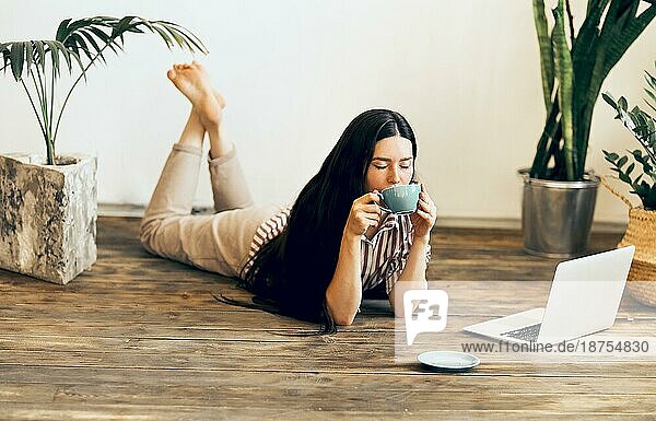 Ruhige Unternehmerin  die zu Hause mit ihrem Laptop auf dem Holzboden liegt und ein heißes Getränk trinkt  während sie aus der Ferne arbeitet