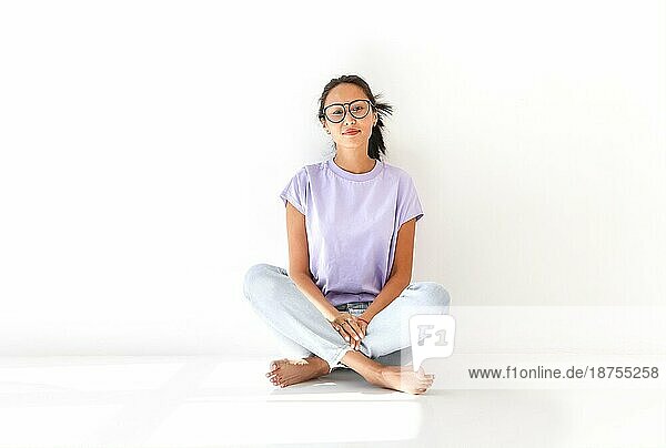 Ganzer Körper einer positiven barfuß tausendjährigen asiatischen Frau in lässigem Hemd und Jeans auf dem Boden sitzend in einem hellen Studio