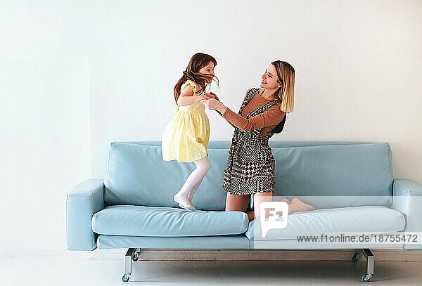Glückliches verspieltes kleines Mädchen Tochter spielt mit positiven Mutter  Springen auf dem Sofa  verbringen Zeit zusammen während der Ferien zu Hause  Mama und Kind Spaß haben im Wohnzimmer