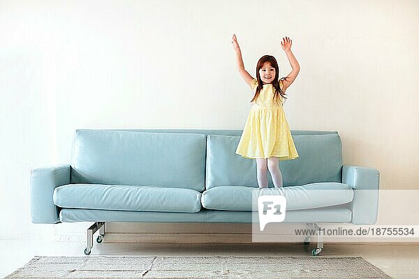 Kleine kaukasische niedlich schönes Mädchen in gelben Kleid spielen und springen auf blaün Sofa im Wohnzimmer zu Hause vor weißem Hintergrundem Betonwand Hintergrund. Kindheit und Freizeit Konzept