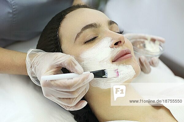 Eine nicht erkennbare Kosmetikerin trägt eine Tonmaske mit einem Pinsel auf das Gesicht einer entspannten Kundin auf  während sie eine Hautpflegeprozedur in einem SpaSalon durchführt