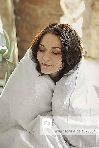 Porträt einer jungen  schönen  glücklichen Frau  die in eine Bettdecke eingewickelt sitzt  die Augen schließt und vor Freude und Komfort lächelt  während sie am frühen sonnigen Morgen in einer gemütlichen  heimeligen Atmosphäre aufwacht