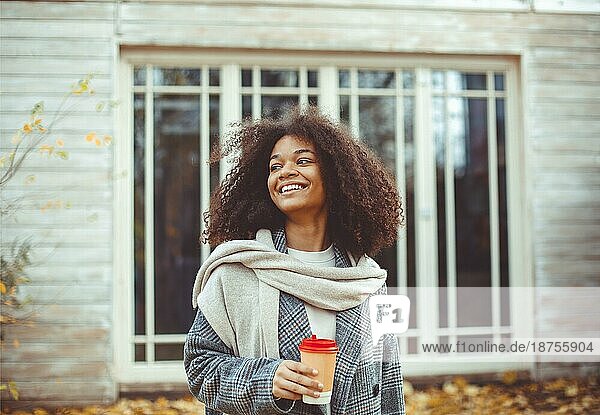 Herbststimmung. Junge glückliche afroamerikanische Frau mit lockigem Haar im Herbst Park hält Kaffee zum Mitnehmen  schaut zur Seite und lächelt  entspannt in der Natur  genießt ihre Lieblingsjahreszeit. Selektiver Fokus