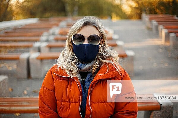 Blonde Frau in Oberbekleidung und Maske geht auf einem gepflasterten Weg an einem sonnigen Tag im Herbstpark während einer Pandemie