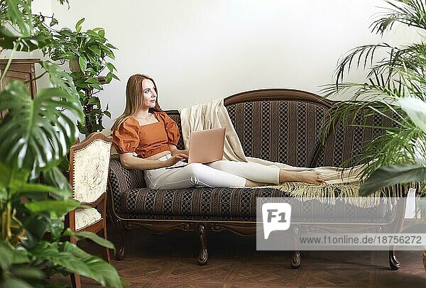 Barfüßige Frau  die ein Netbook benutzt und wegschaut  während sie sich auf einer eleganten Couch inmitten von üppigen Topfpflanzen entspannt