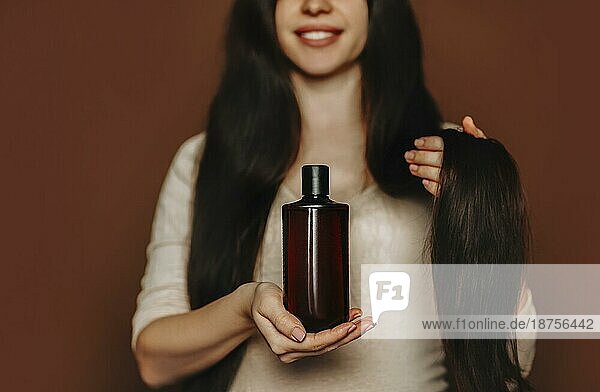 Junge lächelnde Frau mit langen gesunden braunen Haaren  die ein Shampoo mit organischen  natürlichen Inhaltsstoffen in der Hand hält. Glückliche Frau wirbt für Haarpflegeprodukte  während sie vor einem Studiohintergrund steht