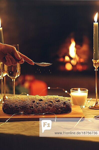 Weihnachtsessen. Weibliche Hand bestreut Puderzucker auf Weihnachten Kuchen Kuchen stehen auf rustikalen Holztisch mit Gläsern Champagner und brennenden Kerzen  vertikale Zusammensetzung. Selektiver Fokus. Stollen