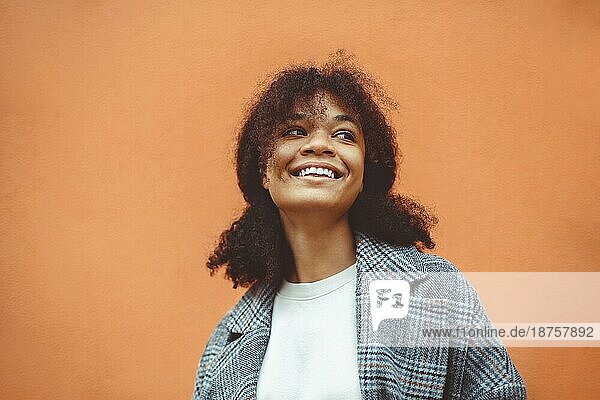 Genießen Sie das Leben. Schöne glückliche afrikanische Ethnizität Mädchen mit üppigen dunklen lockigen Frisur in stilvollen Mantel stehend gegen orange Wand Hintergrund  Blick weg mit breiten hellen Lächeln. Glück Konzept