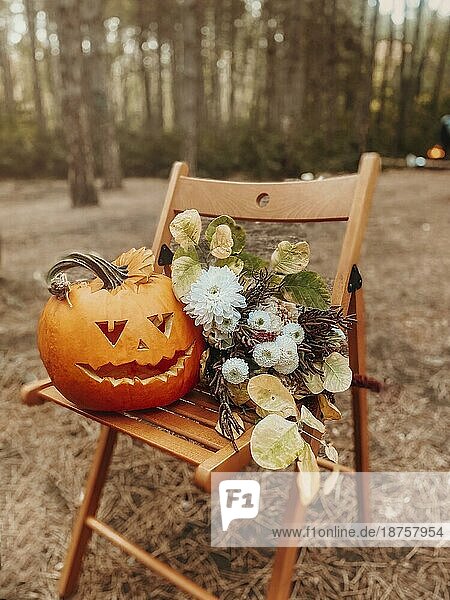 Halloween Dekorationen. Geschnitzte orange Kürbis jack o lantern mit gruseligen Gesicht und Herbst Blumenstrauß auf Holzstuhl im Wald