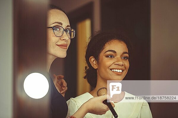 Junge glückliche attraktive afroamerikanische Frau  die einen Visagisten in einem Schönheitssalon besucht  ein professioneller Visagist hält einen Pinsel und trägt Make up auf das Gesicht eines lächelnden schwarzen Mädchens auf  das vor einem Spiegel sitzt