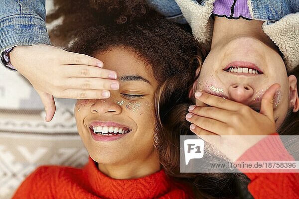 Blick von oben auf zwei glückliche multirassische Teenagermädchen die sich die Augen zuhalten und lächeln  während sie auf einer Decke im Freien liegen und die Jugend und die Gesellschaft des jeweils anderen genießen Diverse weibliche Freundschaft Konzept