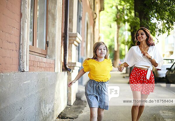Kleines Mädchen hält die Hand einer stilvollen Frau und geht in der Nähe eines alten Gebäudes an einem sonnigen Sommertag auf einer Straße in der Stadt spazieren