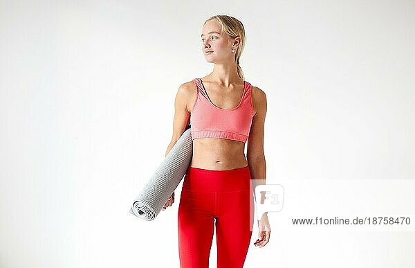 Junge schlanke Frau in leuchtend rotem Oberteil und Leggings hält Matte für Yogaübungen in weißem Studio