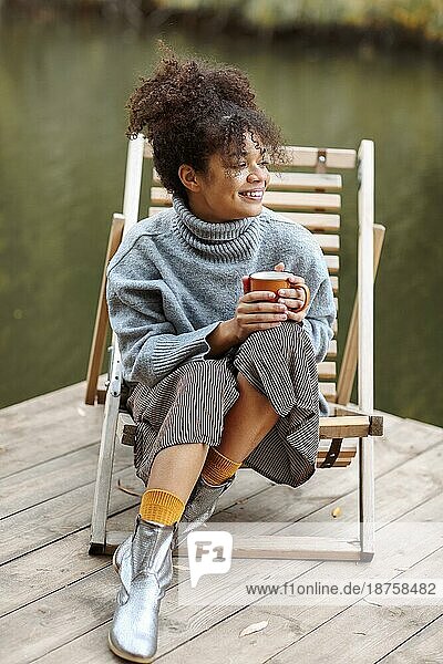 Junge glückliche sorglose afroamerikanische Frau mit einer Tasse heißem Tee  die sich in der Nähe eines Teiches im Herbst entspannt  gemischtrassige Frau im warmen Pullover  die das Herbstwochenende in der Natur genießt. Erholung Konzept