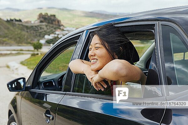 Fröhliche chinesische junge Frau schaut aus dem Autofenster in die Natur