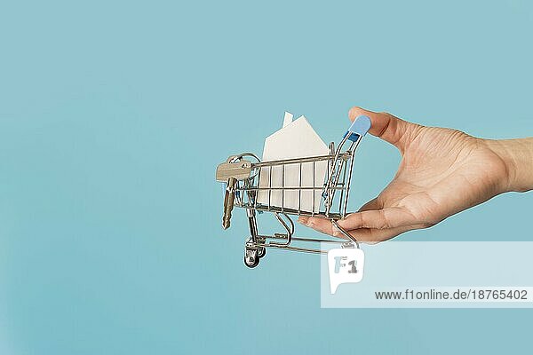 Nahaufnahme Hand hält Miniatur-Einkaufswagen mit Papier Hausschlüssel gegen blaün Hintergrund