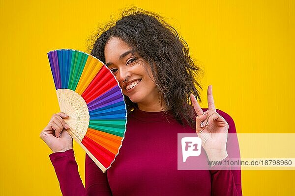 Porträt einer lateinischen Frau lächelnd mit einem Regenbogen lgbt Fan auf einem gelben Hintergrund  so dass der Sieg Symbol