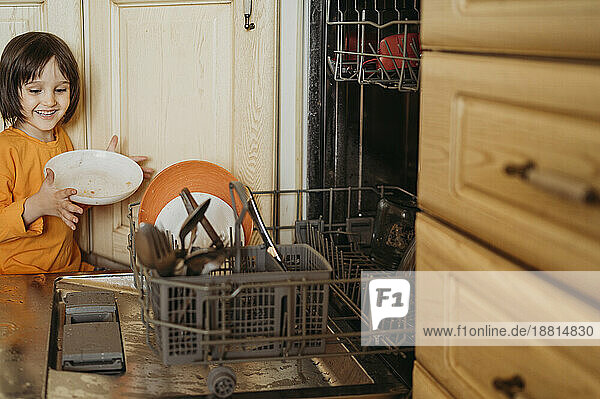 Smiling boy loading plates in dishwasher at home