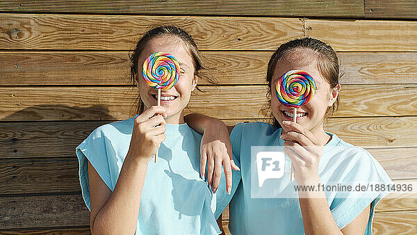 Happy sisters holding lollipop over face in front of wooden wall