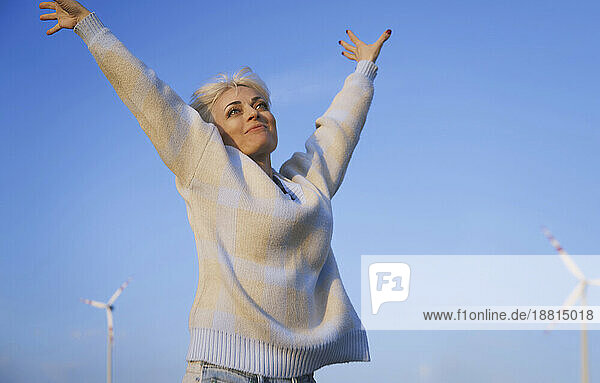 Smiling woman with arms raised standing near wind turbines
