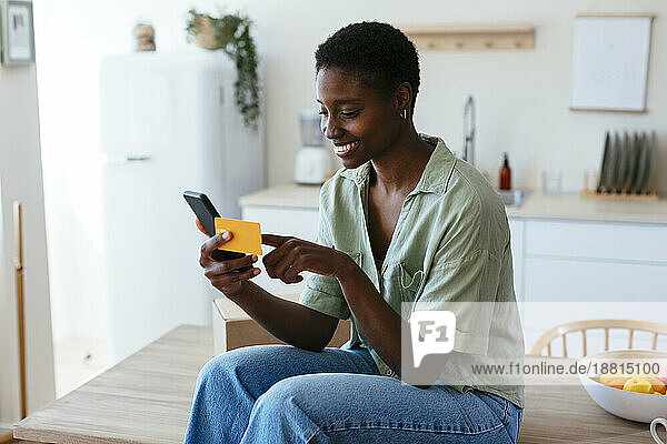 Smiling young woman making payment through credit card on mobile phone at home