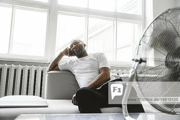 Mann entspannt sich zu Hause auf Sofa vor elektrischem Ventilator