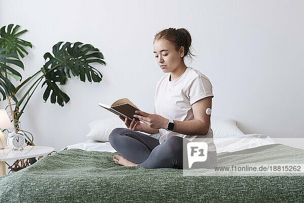 Woman with diabetes reading book sitting cross-legged on bed at home