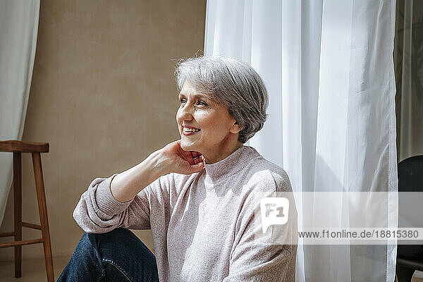 Thoughtful smiling woman sitting by curtain at home