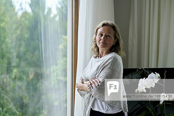 Thoughtful senior woman standing with arms crossed by window at home