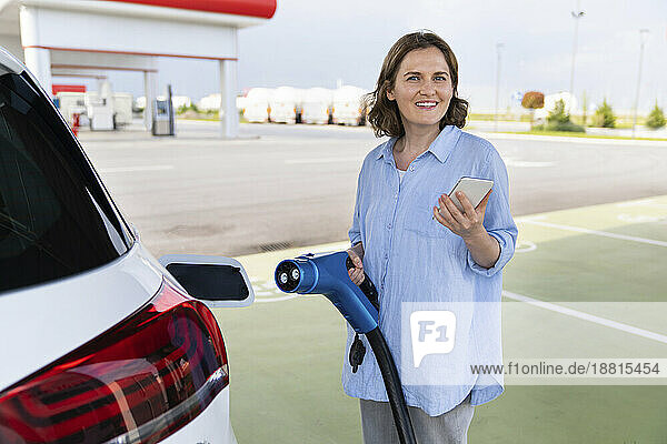 Frau hält Autoladegerät und Smartphone in der Nähe eines Elektroautos am Bahnhof