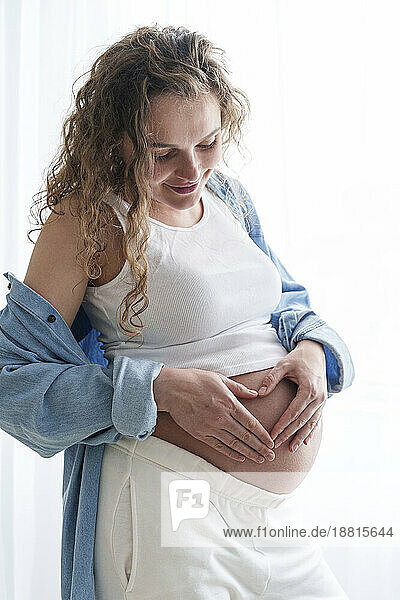 Lächelnde schwangere Frau  die am Fenster eine Herzform auf dem Bauch macht