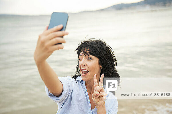 Frau macht ein Friedenszeichen und macht ein Selfie mit ihrem Smartphone am Strand