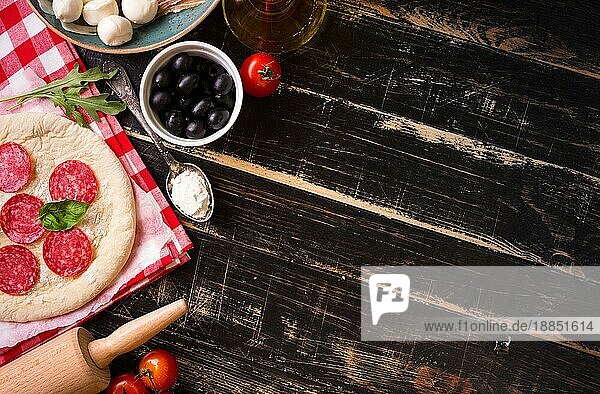 Hintergrund zum Pizzabacken. Zutaten für die Herstellung von Pizza. Platz für Text. Pizzateig  Mehl  Käse  Mozzarella  Tomaten  Basilikum  Peperoni  Oliven und Nudelholz auf schwarzem Holzhintergrund. Ansicht von oben