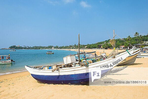 Am Strand eines der wichtigsten Touristenzentren im Südwesten Sri Lankas. Touristen nehmen ein Sonnenbad und treiben Wassersport. Auslegerboote und traditionelle Fischerboote am Strand und vor Anker